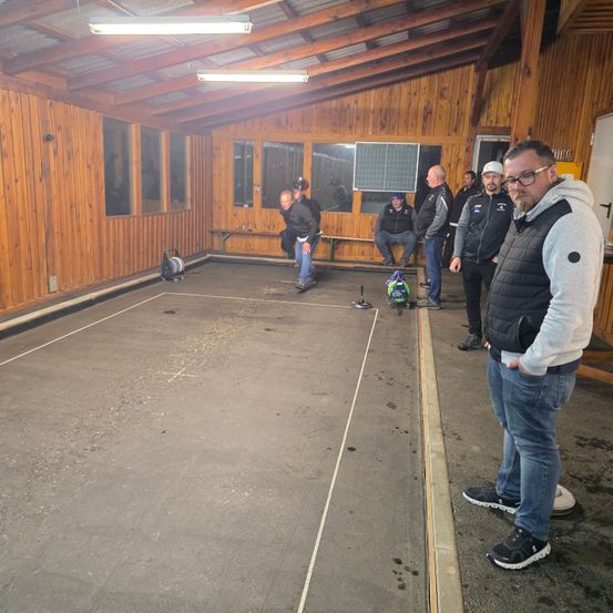 Eine Gruppe von Menschen in einem Raum mit Holzwänden und Bänken, ein Mann beugt sich vor, während ein anderer steht und zusieht.