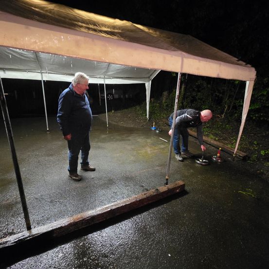 Zwei Männer spielen unter einem Zelt auf einer nassen Oberfläche ein Curlingspiel. Ein Mann hält einen Besen, während der andere steht und zuschaut.