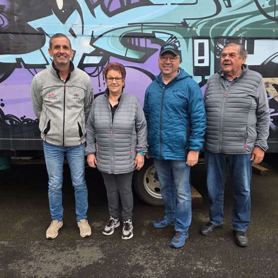 Vier Menschen, zwei Männer und zwei Frauen, stehen vor einem mit Graffiti bedeckten LKW. Sie alle lächeln und schauen zur Kamera.