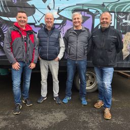 Vier Männer stehen zusammen vor einer farbenfrohen, mit Graffiti bedeckten Wand. Sie alle lächeln und scheinen für ein Foto zu posieren.