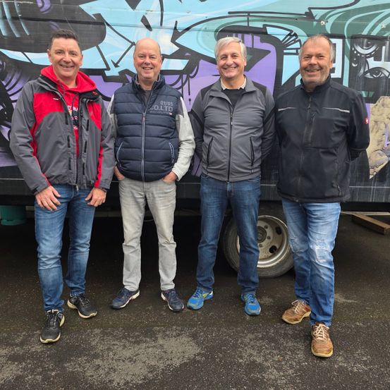 Vier Männer stehen zusammen vor einer farbenfrohen, mit Graffiti bedeckten Wand. Sie alle lächeln und scheinen für ein Foto zu posieren.