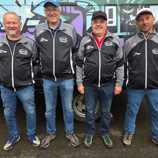 Vier Männer in schwarzen und grauen Jacken mit weißen Streifen stehen vor einem mit Graffiti bedeckten Fahrzeug. Sie alle lächeln und scheinen für ein Foto zu posieren. Der Mann in der Mitte hat ein rotes Innenhemd.