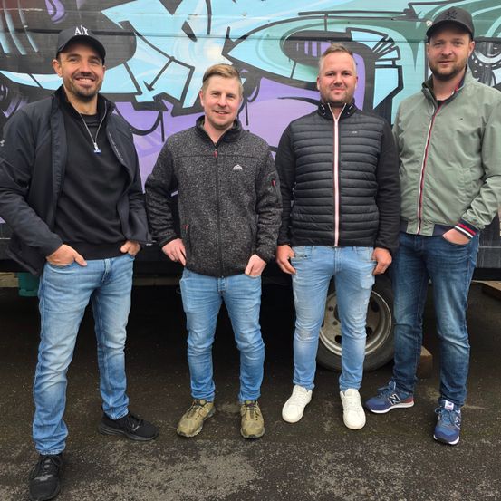 Vier Männer, die nebeneinander stehen, die Hände in den Taschen, lächeln, mit einem Graffiti-Truck im Hintergrund.
