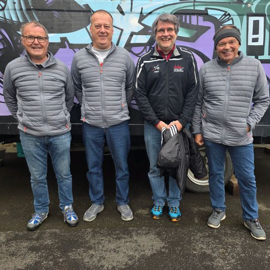 Vier Männer stehen vor einem bunten Graffiti-Truck, lächeln und sind in Freizeitkleidung gekleidet. Der Mann in der Mitte hält eine schwarze Jacke.