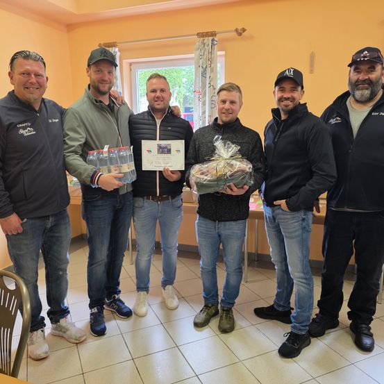 Fünf Männer stehen in einem Raum, lächeln und posieren für ein Foto. Sie tragen alle Jacken und Turnschuhe. Der Mann in der Mitte hält ein Zertifikat und einen Geschenkkorb. Der Mann auf der linken Seite hält eine Flasche Wasser. Hinter ihnen steht ein Tisch mit einem Stuhl und einem Fenster mit Vorhängen.