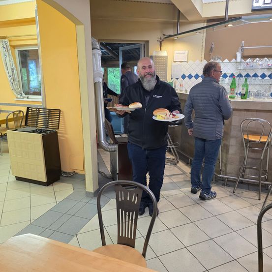 Ein Mann in einer schwarzen Jacke hält zwei Teller mit Essen in einem Restaurant. Ein weiterer Mann in einer grauen Jacke steht hinter ihm, und im Vordergrund befindet sich ein Tisch mit Stühlen.