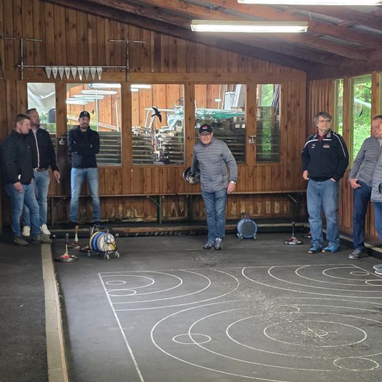 Mehrere Männer stehen in einem hölzernen Raum mit weißen Markierungen auf dem Boden. Zwei Männer halten einen Croquetschläger und ein anderer Mann ein Helm.