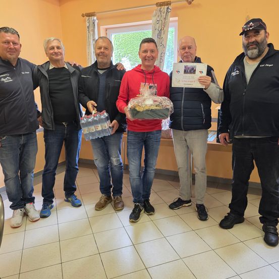 Sechs Männer stehen in einer Reihe in einem Raum, möglicherweise einem Besprechungsraum, und tragen Jacken. Ein Mann hält ein Zertifikat, während ein anderer einen Geschenkkorb hält.