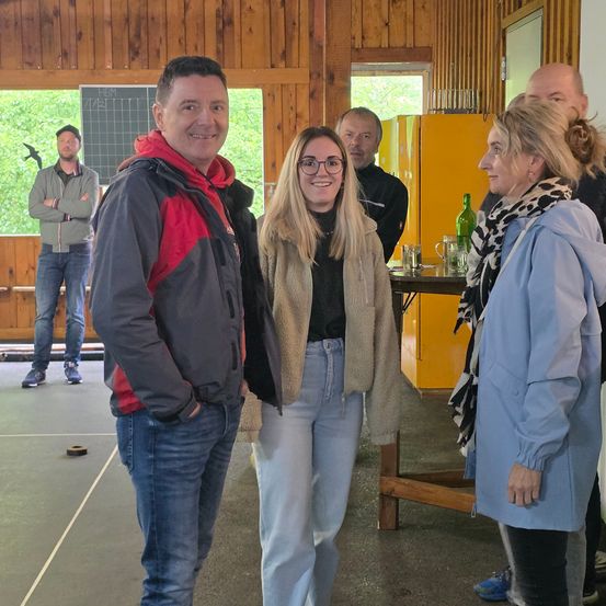 Drei Menschen stehen in einem Raum und unterhalten sich. Hinter ihnen steht eine Person in der Nähe des Glasfensters. Ein Holztisch steht in der Nähe des Fensters, und eine Flasche darauf.