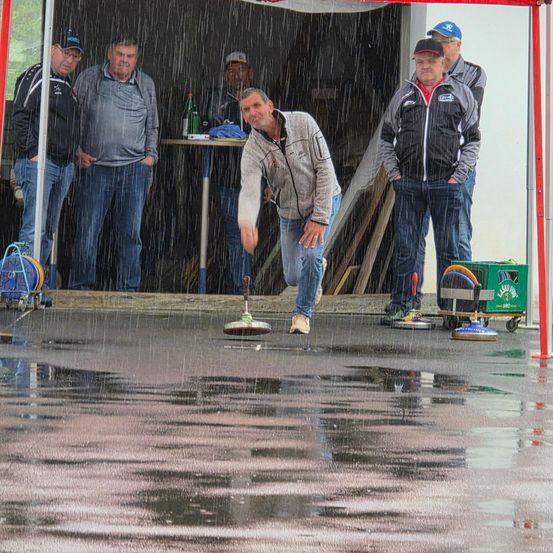 Ein Mann in einer grauen Jacke wirft unter starkem Regen einen Curling-Stein in einem Außenbereich. Andere Männer stehen im Hintergrund in einem überdachten Bereich.