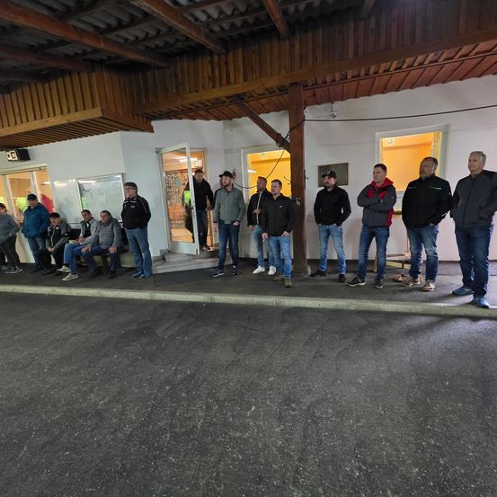 Eine Gruppe von Menschen in Jacken steht vor einem Gebäude. Einige sitzen auf einer Bank in der Nähe eines Eingangs.