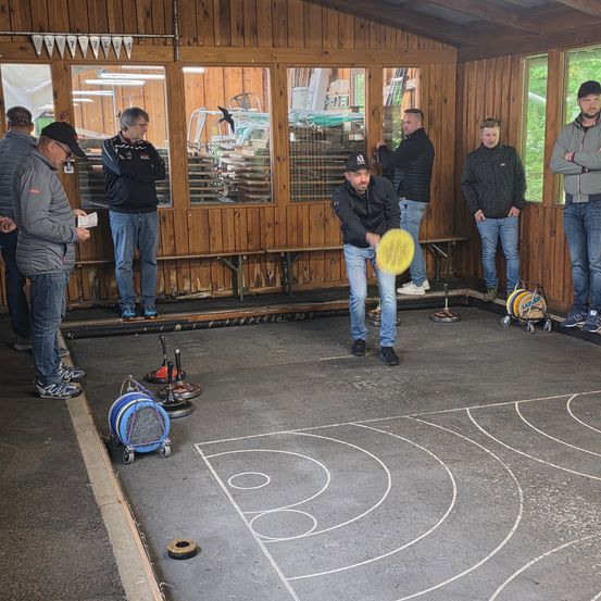 Eine Gruppe von Menschen versammelt sich in einem Raum mit gelbem Boden. Ein Mann wirft einen gelben Frisbee.