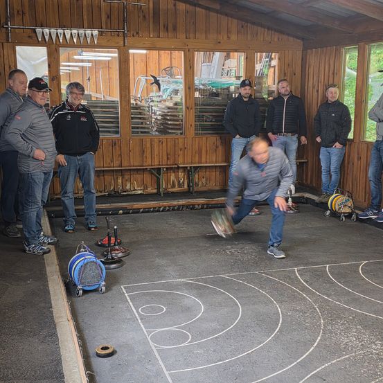 Eine Gruppe von Männern versammelt sich in einem Raum mit Holzwänden und Glasfenstern. Ein Mann spielt ein Spiel auf einem markierten Boden, während andere zusehen.