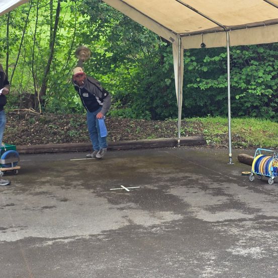Ein Mann mit Hut und einer Zigarette im Mund wirft einen Frisbee unter einem Zelt auf einem Parkplatz.