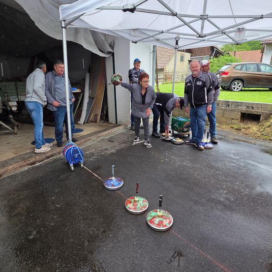 Eine Gruppe von Menschen steht unter einem Zelt in einer Garage, eine Frau wirft einen Ball. Der Boden ist gepflastert und darauf befinden sich Gegenstände. Dahinter ist ein Auto auf dem Rasen geparkt.