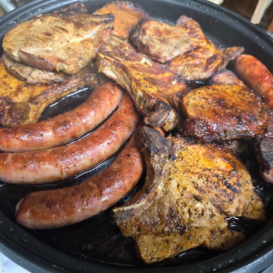 Nahaufnahme verschiedener Fleischsorten in einer Grillpfanne, darunter Schweinekoteletts, Rippchen, Würste und anderes Fleisch, alle mit verkohlter und rauchiger Erscheinung.