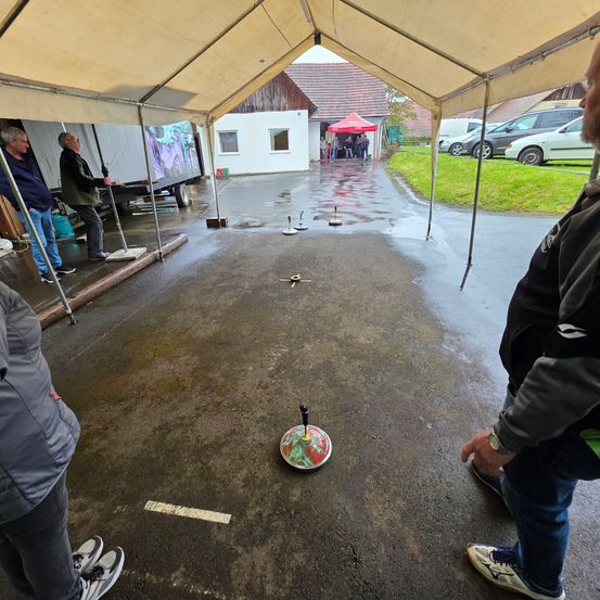 Mehrere Personen stehen unter einem Zelt, wobei eine Person im Vordergrund einen sich drehenden Top betrachtet.