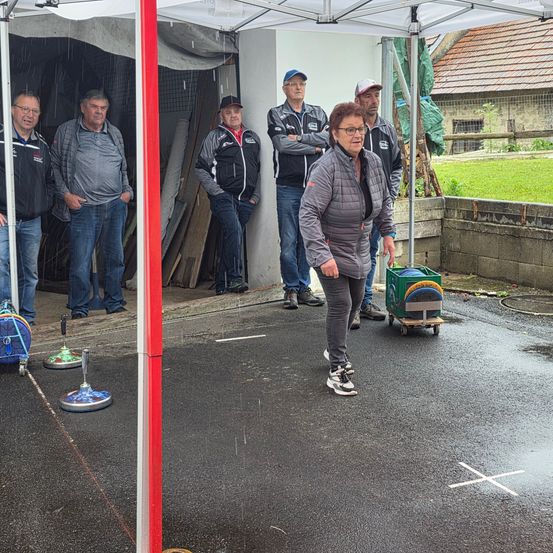 Eine Frau geht auf einen Pfosten zu, während andere zusehen. Zwei Pfosten mit Bürsten stehen auf dem Boden. Ein Trolley steht hinter ihnen, und ein Gebäude ist im Hintergrund.