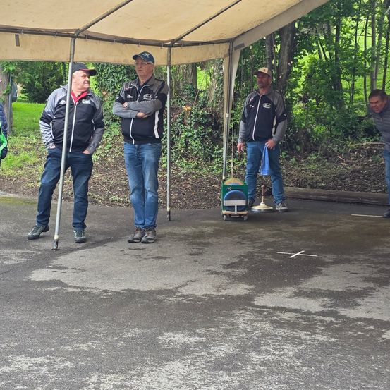 Drei Männer stehen unter einem Zelt auf einer gepflasterten Fläche mit Grünpflanzen im Hintergrund. Ein Mann hält einen kleinen Trolley.