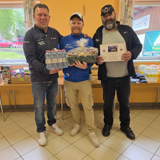 Drei Männer stehen zusammen. Einer hält eine Wasserflasche, ein anderer einen Korb mit Essen und der dritte ein Zertifikat. Sie befinden sich in einem Raum mit einem Tisch, Kisten und Flaschen im Hintergrund.