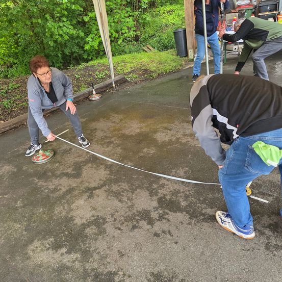 Zwei Personen spielen ein Spiel auf einer Betonfläche. Eine Frau mit Brille hält einen Stock und eine runde Scheibe. Ein Mann beugt sich vor, scheinbar konzentriert auf das Spiel.