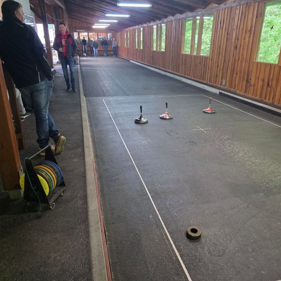 Eine Bowlingbahn mit drei Bowlingpins auf dem Boden. Ein Mann steht neben einem Ständer mit Bowlingbällen.