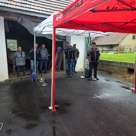 Eine Gruppe von Menschen steht unter einem roten Zelt auf einer nassen Straße, ein Mann hält einen Helm, während andere in Jacken und Turnschuhen gekleidet sind. Ein Rasen und ein Gebäude sind im Hintergrund zu sehen.