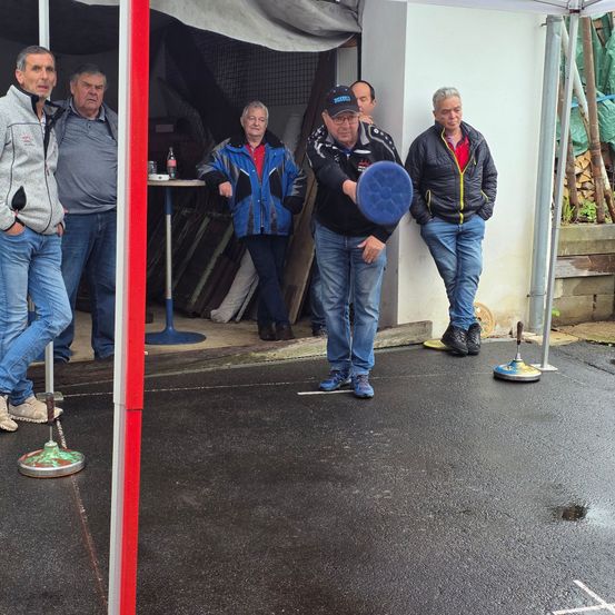 Männer spielen Boule unter einem Zelt, einer wirft einen blauen Ball, während die anderen zusehen.