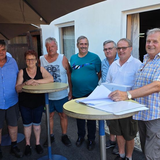 Eine Gruppe von Menschen steht vor einem Gebäude. Ein Mann hält eine Schachtel und Papiere. Vor ihnen stehen zwei Tische. Der Mann in der Mitte lächelt und schaut zur Kamera.