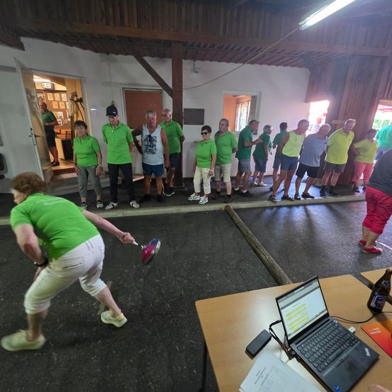 Eine Gruppe von Menschen in grünen Shirts versammelt sich in einem Raum, eine Frau spielt Tischtennis. Ein Tisch mit Laptop und Flasche befindet sich in der Nähe.