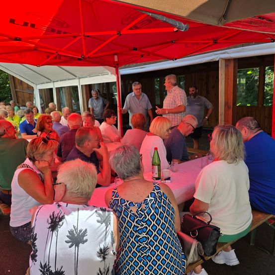 Eine Gruppe älterer Menschen sitzt unter einem roten Baldachin um einen Tisch. Ein Mann steht und spricht zu ihnen. Sie befinden sich in einem Außenbereich, wahrscheinlich einem Restaurant.
