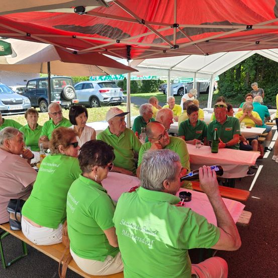 Eine Gruppe älterer Erwachsener in grünen Shirts sitzt unter einem roten Zelt und trinkt Bier. Einige tragen Sonnenbrillen und Hüte. Im Hintergrund sind Autos geparkt.