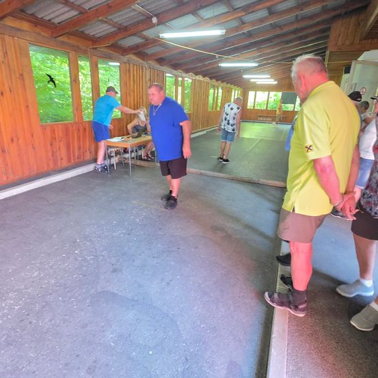 Eine Gruppe von Menschen befindet sich in einem holzgetäfelten Raum. Zwei Männer gehen auf einen Tisch zu, an dem ein anderer Mann steht. Ein Vogel fliegt vor dem Fenster.