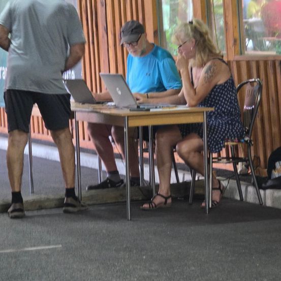 Drei Personen sitzen um einen Tisch herum, zwei benutzen Laptops, eine Frau beobachtet und ein Mann steht in der Nähe. Die Umgebung ist im Freien mit Holzwänden und Blick auf ein Auto draußen.