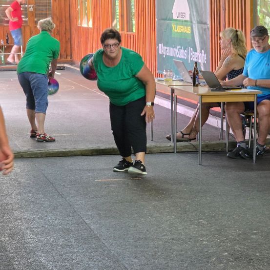 Eine Gruppe von Menschen spielt draußen Boule. Eine Frau wirft einen Ball, während andere zuschauen. Zwei Personen sitzen auf Stühlen an einem Tisch mit Laptops und einer Bierflasche.