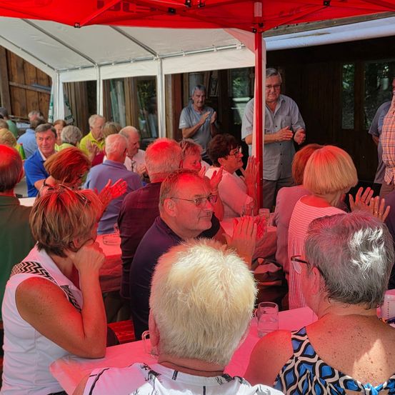 Eine Gruppe älterer Erwachsener versammelt sich unter einem roten Zelt. Einige klatschen, während andere sitzen und reden. Zwei Männer stehen in der Nähe des Eingangs, und eine Frau ist teilweise im Hintergrund zu sehen.