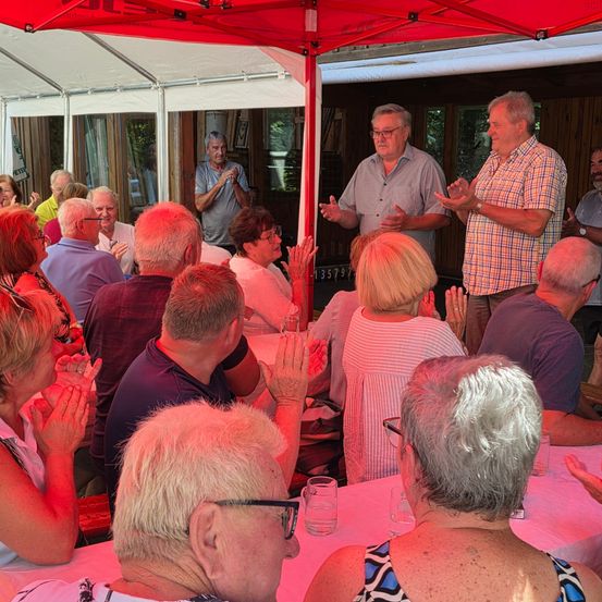 Eine Gruppe von Menschen klatscht und beobachtet zwei Männer, die unter einem roten Zelt stehen. Die Männer lächeln und gestikulieren. Eine Frau in einem weißen Shirt klatscht. Auf dem Tisch stehen Gläser und Teller.