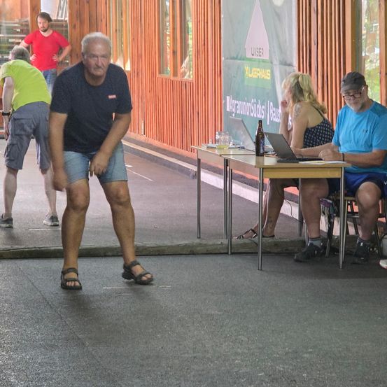 Eine Gruppe von Menschen ist in einem hölzernen Gebäude versammelt. Ein älterer Mann geht auf die Kamera zu. Ein Tisch mit Laptop und Flasche steht hinter einem sitzenden Mann und einer Frau.