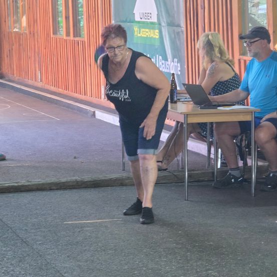 Eine ältere Frau steht in einer lässigen Kleidung und scheint auf einen Tisch mit einem Laptop und einer Flasche zuzugehen. Zwei Personen sitzen am Tisch, eine mit einer Brille, die andere mit einer Mütze. Eine Holzwand mit einem Schild ist im Hintergrund.