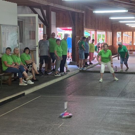 Eine Gruppe von Menschen in grünen Shirts beobachtet eine Frau beim Shuffleboard-Spiel. Der Shuffleboard-Platz hat einen nassen Boden.