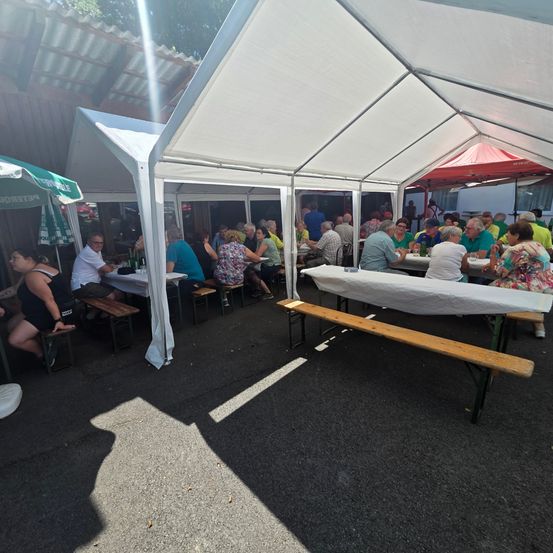 Eine Gruppe von Menschen sitzt an Tischen unter einem weißen Zelt, mit mehreren Zelten im Hintergrund. Einige Tische sind mit weißen Tüchern bedeckt, und der Bereich ist schattig.