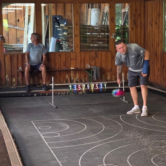 Zwei Männer spielen eine Partie Boccia. Ein Mann beobachtet vom Holzbank, während der andere einen Ball wirft.