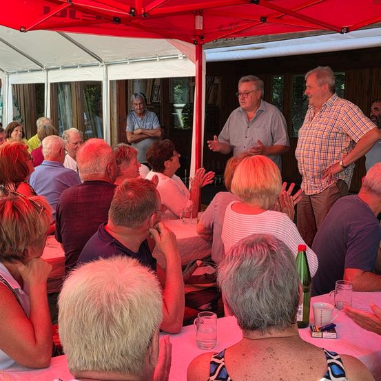 Eine Gruppe älterer Menschen sitzt unter einem roten Zelt um Tische herum und hört zwei Männern zu, die sprechen. Ein Mann gestikuliert, während der andere zuhört. Gläser, eine Flasche und eine Tasse stehen auf dem Tisch.