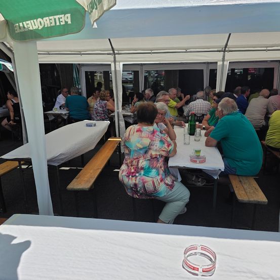 Mehrere Personen sitzen unter einem Zelt an Tischen und unterhalten sich. Das Zelt hat ein weißes Dach mit Text, und die Tische sind mit weißem Tuch bedeckt. Flaschen, Gläser und Aschenbecher stehen auf den Tischen.