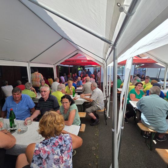 Eine Gruppe von Menschen sitzt unter einem Zelt an Tischen. Einige Tische haben Flaschen und Gläser. Das Wetter ist sonnig.