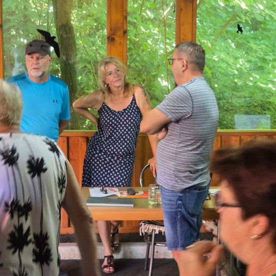 Eine Gruppe von Menschen ist in einem Raum mit Holzwänden und Glasfenstern. Zwei Frauen und zwei Männer unterhalten sich. Eine Frau in einem blauen Kleid steht und schaut den Mann mit Brille an. Ein anderer Mann steht in der Nähe des Glasfensters. Ein Tisch hat Papiere, eine Flasche und andere Gegenstände darauf.