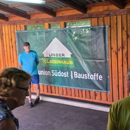 Ein Mann in einem blauen Shirt und eine Frau mit Brille stehen vor einem Holzgebäude mit einem grünen Banner.