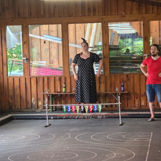 Eine Frau in einem gepunkteten Kleid steht auf einer Bühne mit einer Nummernspur, während ein Mann in einem roten T-Shirt neben ihr steht. Hinter ihnen spiegeln Holzwände und Glasfenster die Außengrüne wider.