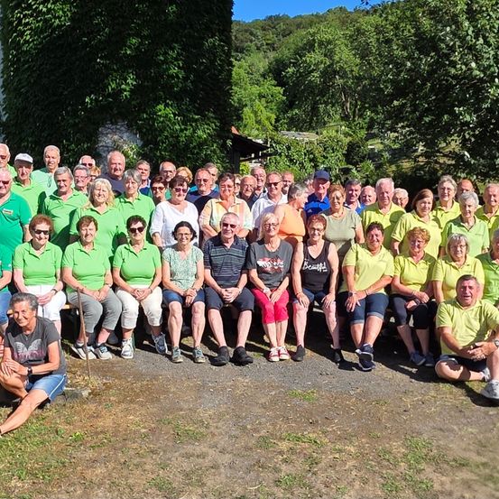 Eine Gruppe von Menschen, die meistens grüne Hemden tragen, sind im Freien versammelt. Sie sitzen und stehen vor einer Kulisse aus Bäumen und Grün. Einige Personen tragen Hüte und Sonnenbrillen.