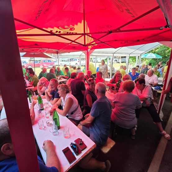 Eine Gruppe von Menschen, einige in Grün, sitzen unter einem roten Zelt um Tische herum, mit Flaschen, Gläsern und Telefonen auf dem Tisch. Im Hintergrund sitzen weitere Menschen, und Autos sind geparkt.